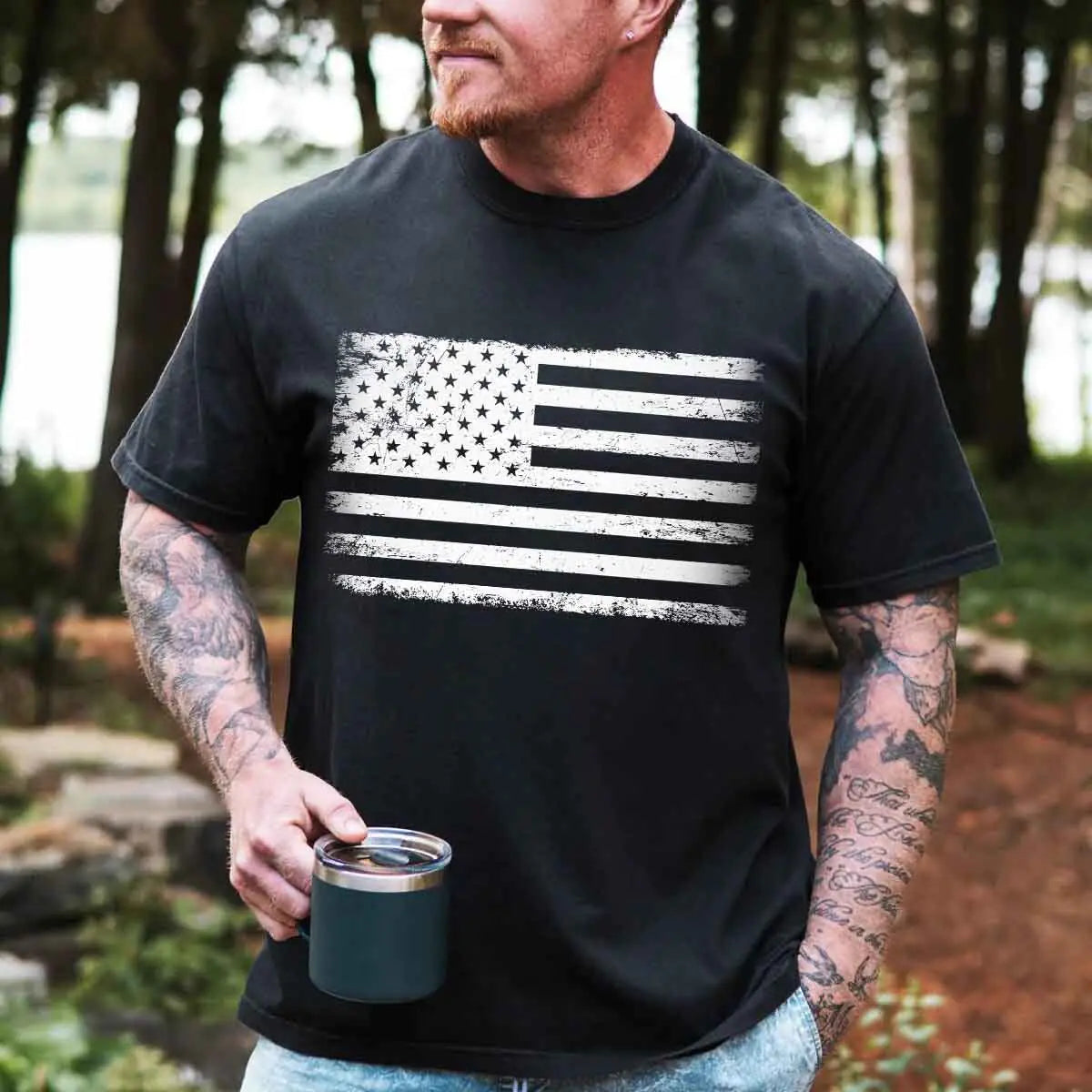 Man wearing a black t-shirt with an American flag design, holding a mug outdoors.