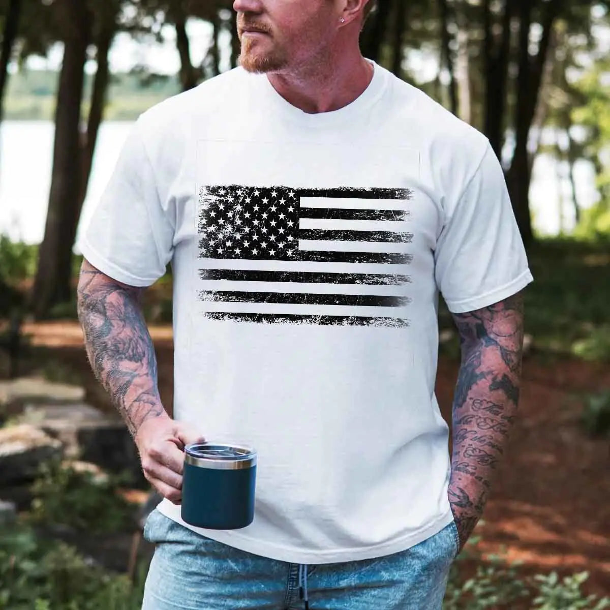 Man wearing a white t-shirt with an American flag design, holding a blue mug outdoors.